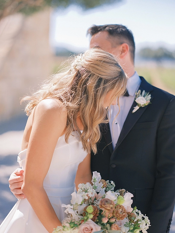 Couple portrait of bride leaning on groom, embracing as she looks down, holding a white blush bouquet outdoors by a stone column