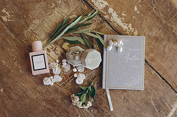 Wedding details flatlay with wedding vow book, rings in a velvet box, perfume, pearl earrings, and greenery on a rustic wooden surface