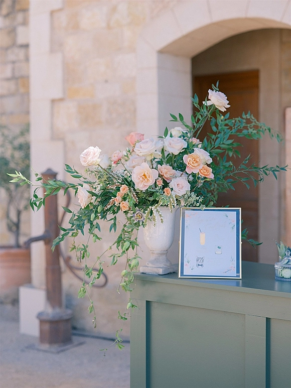 Welcome table decor with a framed sign and urn of blush and peach roses on a green cabinet at a stone-arched outdoor entryway