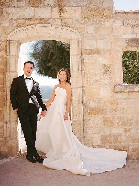 Couple portrait of bride and groom holding hands, her strapless gown with long train beside his black tuxedo under a stone archway
