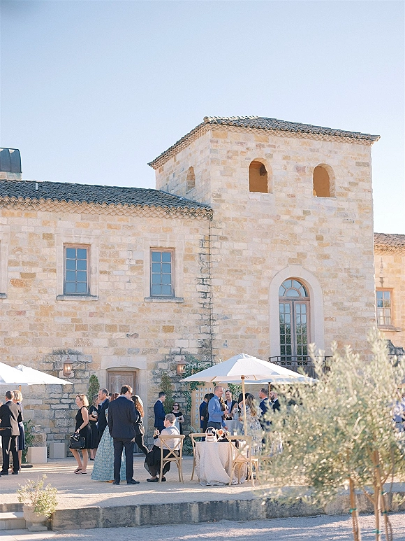 Cocktail hour setup with bistro tables, linens and cocktails under patio umbrellas in a stone courtyard with arched windows and olive trees