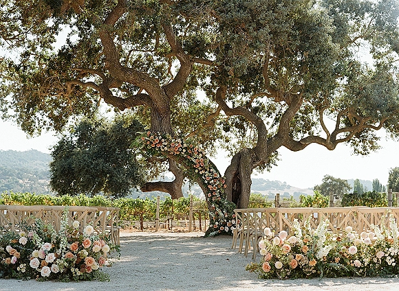 Ceremony setup for an outdoor wedding ceremony with a floral arch and rose aisle arrangements beneath oak trees in a vineyard backdrop