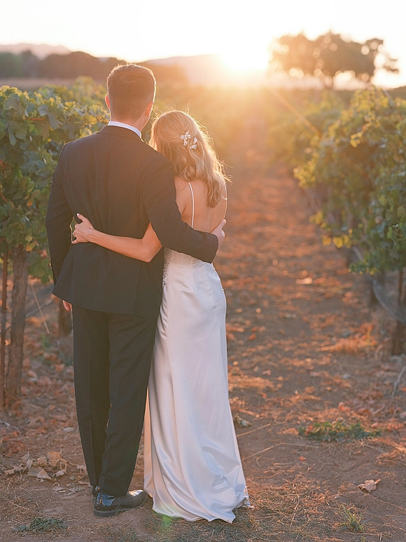Couple portrait of bride and groom from behind walking down a vineyard dirt path at sunset, her low-back dress and hair clip visible