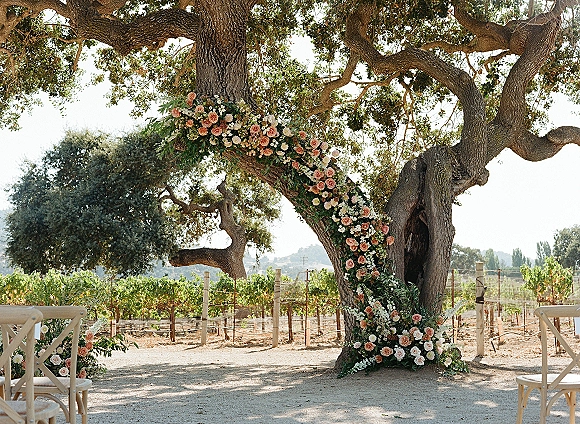 Ceremony altar decor with tree ceremony backdrop, featuring a curved rose-and-greenery floral installation beneath a large oak tree in a vineyard setting