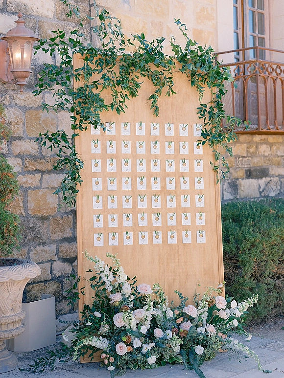 Wedding escort display with an escort card wall on a wood board, framed by greenery garland and blush florals against a stone wall patio backdrop