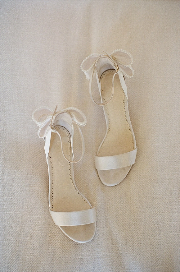Bridal shoes in a wedding shoe flat lay, white ankle-strap heels with bow ties and pearl trim on textured fabric surface