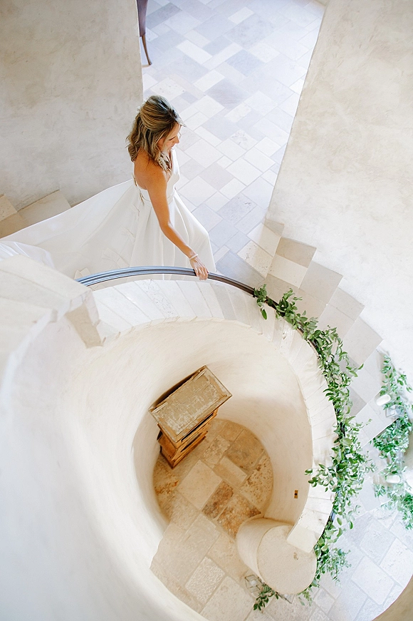 Bridal portrait of a bride on staircase, shot overhead as she holds the railing, her long wedding dress train on stone spiral steps with greenery garland