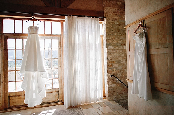 Wedding dresses hanging on dress hangers by sheer curtains, showing strapless and off-the-shoulder lace bodices in window light