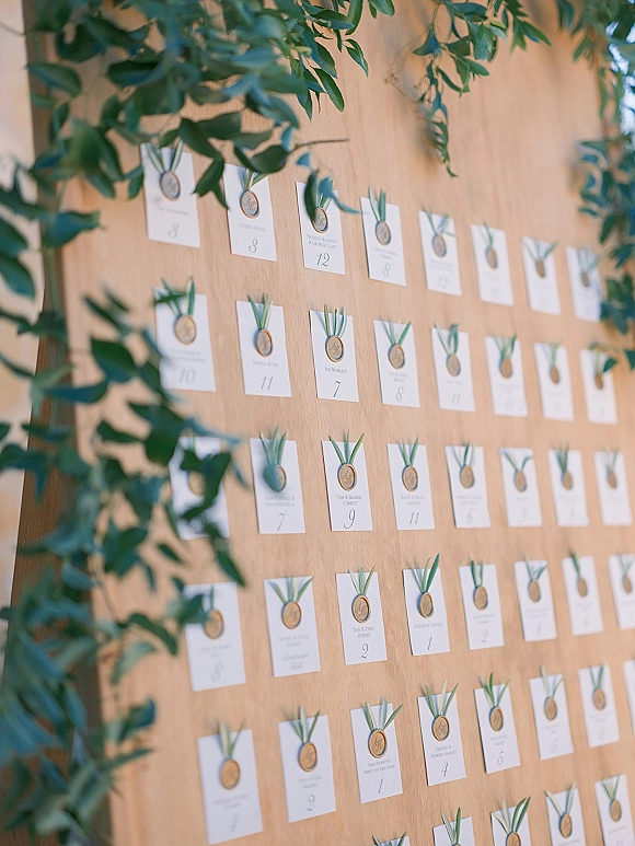 Wedding seating chart with white place cards mounted on a wood board, finished with gold wax seals and ribbon, accented by greenery