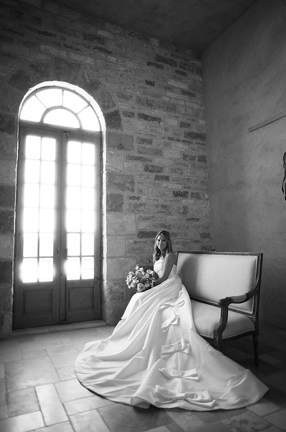 Bridal portrait of a bride sitting on a settee in a strapless gown with long train, holding bouquet, by arched glass door in stone room