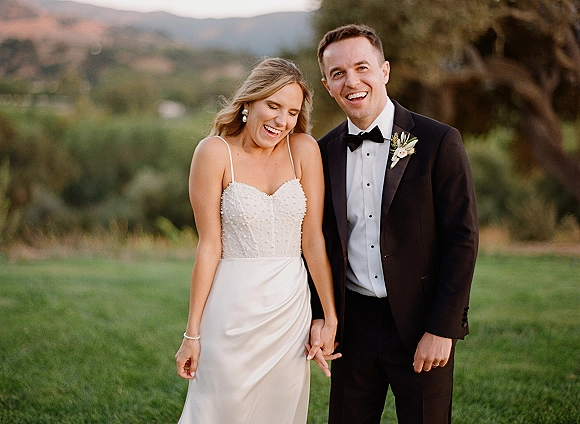 Couple portrait of bride and groom laughing while holding hands, her strapless dress and his tuxedo glowing at sunset in a grassy field