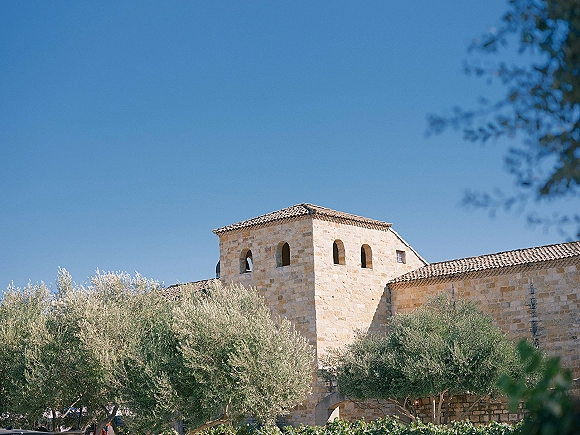 Stone church exterior with terracotta roof tiles, arched windows, and olive trees under a bright blue sky and lush greenery