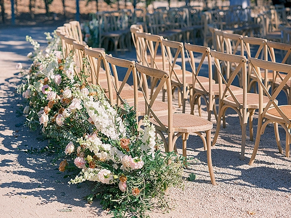 Ceremony aisle decor with blush and white rose ground florals and greenery bordering a gravel path between wood crossback chairs in sunlit trees