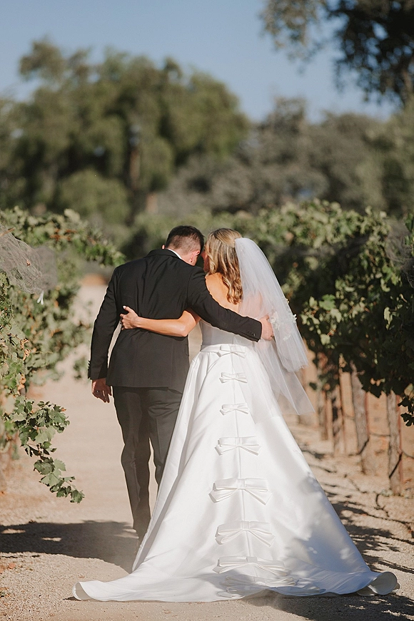 Couple portrait of bride and groom walking away arm in arm, her bow-back gown and veil flowing along a vineyard dirt path