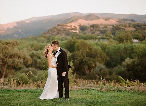Wedding kiss as bride and groom kissing in a mountain landscape, her slip dress and his black tuxedo under string lights at sunset
