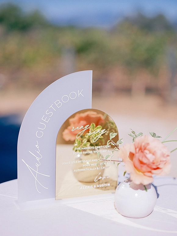Wedding guestbook sign, acrylic guestbook sign in arched clear acrylic with calligraphy on a white tablecloth, bud vase with peach rose and greenery, blurred hills behind