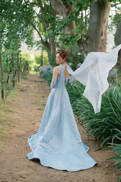 Bridal portrait of a bride in a light blue wedding dress with a sheer cape, along a tree-lined garden path dotted with purple flowers.
