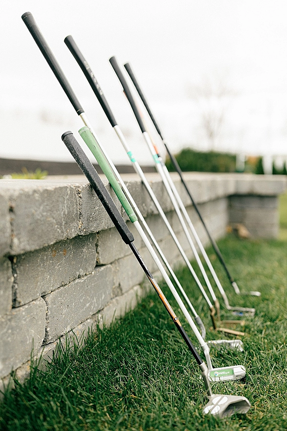 Wedding golf putters lined up for wedding mini golf on a grassy lawn beside a stone retaining wall under open sky
