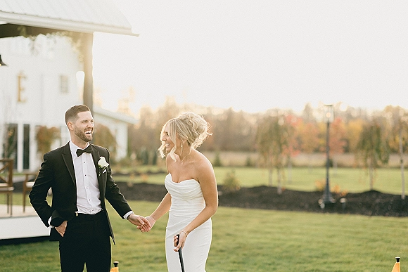 Couple portrait of bride and groom laughing and holding hands at sunset on a lawn near a farmhouse, bride in strapless gown