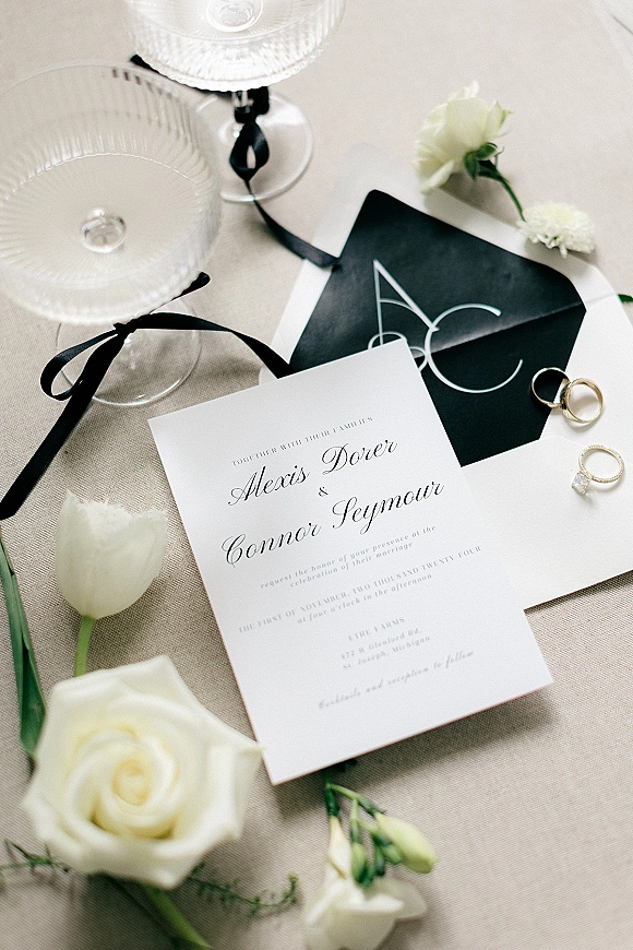 Invitation suite with black and white wedding invitations styled on linen fabric, with black ribbon and gold wedding bands beside champagne coupes