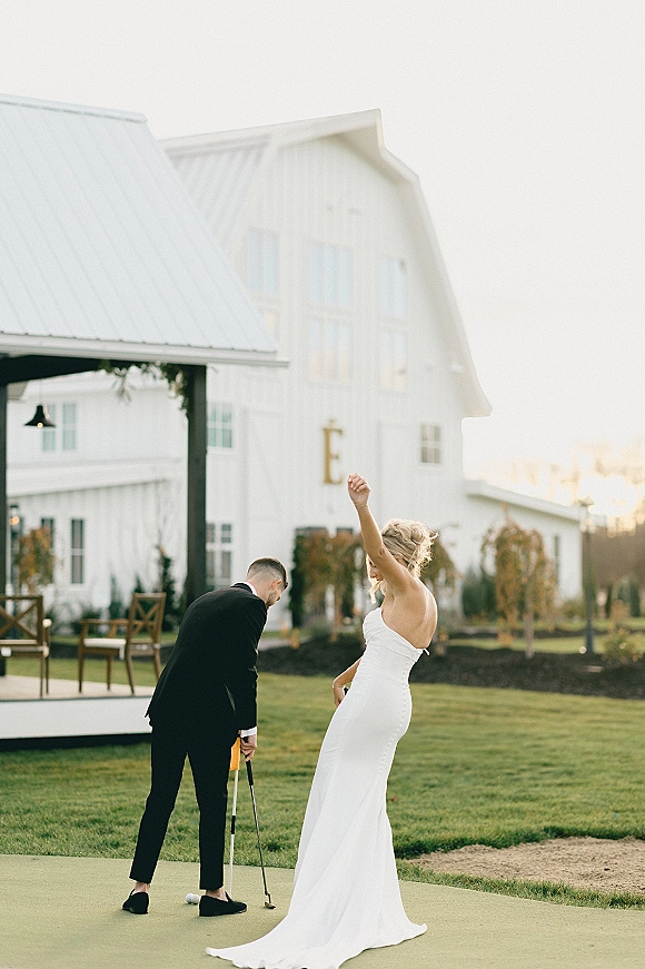 Couple portrait of bride and groom golfing with a putter and golf ball, her strapless dress and his tux in front of a white barn