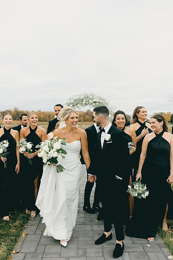 Wedding party portrait of bride and groom walking with bridesmaids and groomsmen, bride holding white bouquet near a floral arch outdoors