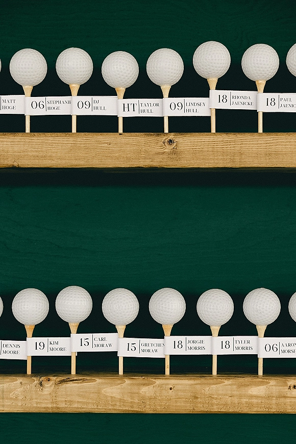 Escort card display with golf themed escort cards on wooden shelves, featuring golf balls, tees, and name tags against a green wall backdrop