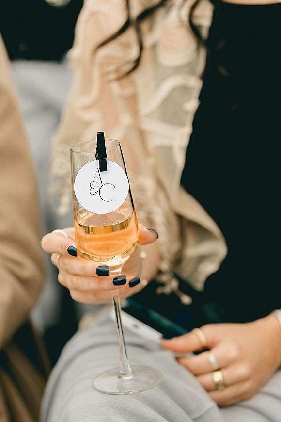 Wedding champagne toast with rosé champagne flute close up, monogram drink tag on mini clothespin, rings and black manicure in blurred reception background
