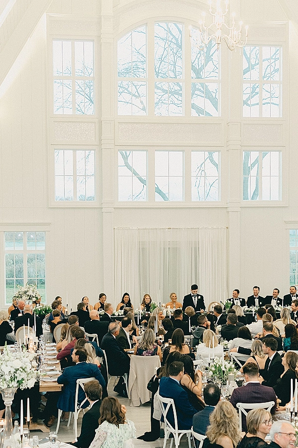 Wedding reception speech during a best man toast at the head table, with taper candles, white florals, chandelier, and bright windows behind