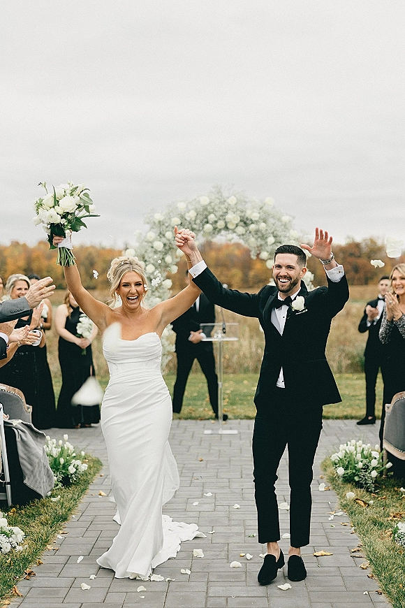 Wedding recessional as bride and groom cheering, holding hands down the aisle with rose petal accents in an outdoor autumn field ceremony