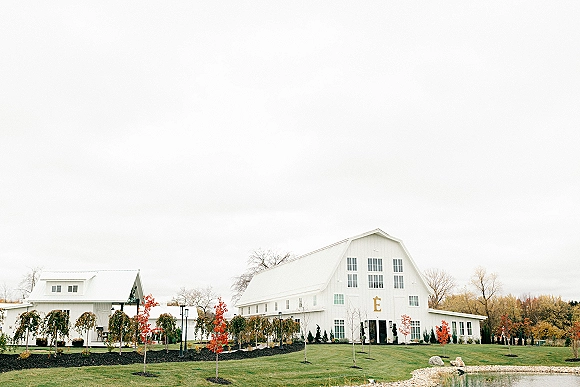 Wedding venue exterior of a white barn building with large windows, lamppost-lined walkway and landscaping rocks beside a pond under cloudy sky
