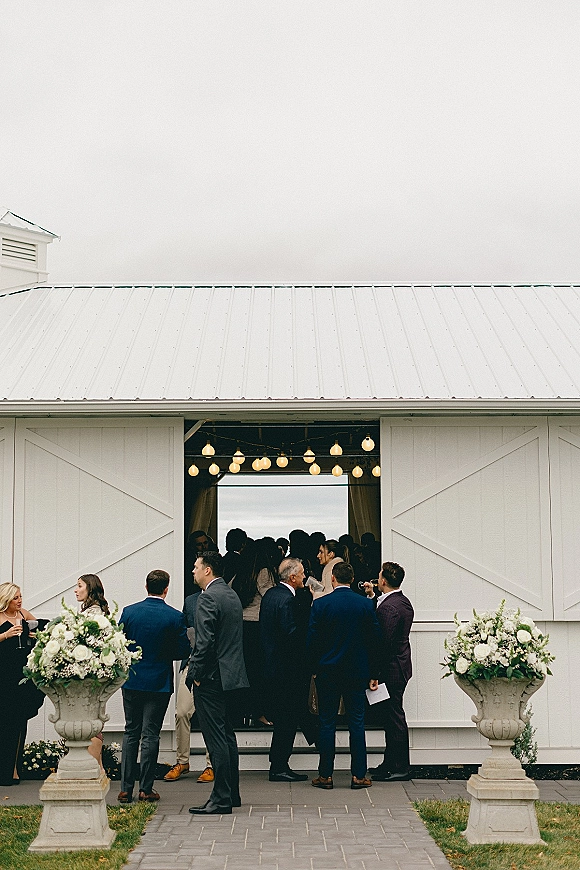 Wedding cocktail hour with wedding guests mingling, holding drinks by white barn doors under string lights and white rose urn florals