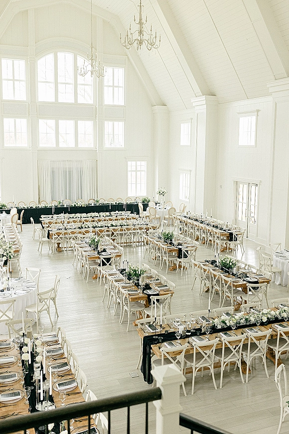 Reception tablescape with wedding banquet table setup featuring long tables, greenery garlands, floral centerpieces, and candles in a bright white hall with chandeliers