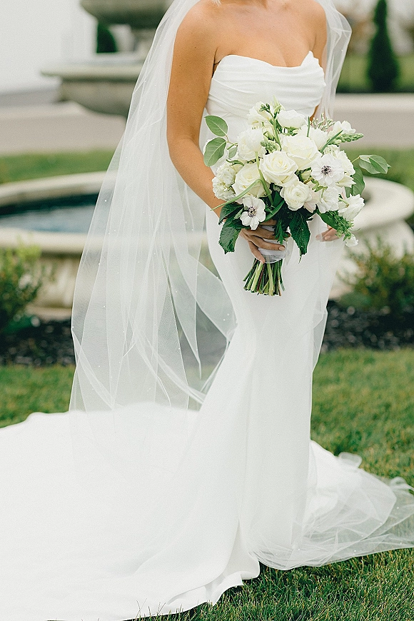 Bridal bouquet of white roses and anemones with greenery held by a bride in a strapless dress on a garden lawn by a stone fountain