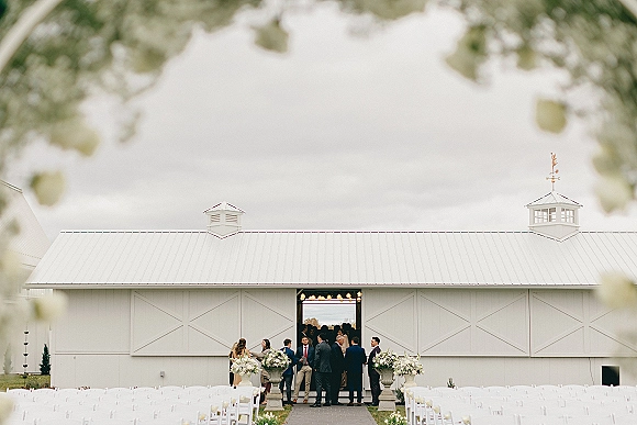 Ceremony setup with outdoor ceremony aisle, white folding chairs and aisle runner leading to barn doors with floral urns and string lights