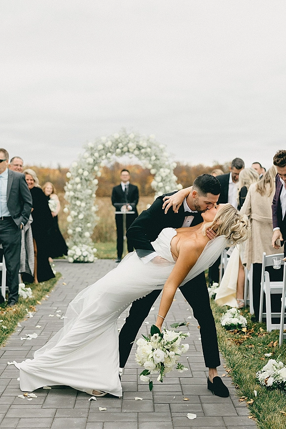 Wedding kiss portrait of bride and groom in a romantic dip, veil flowing, white bouquet, under a floral arch on a stone aisle outdoors.