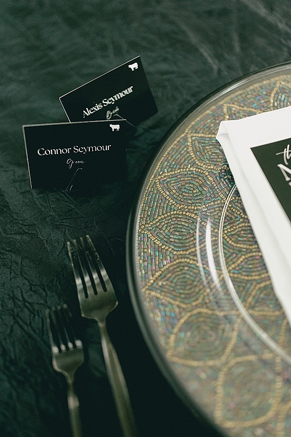 Wedding place cards in black with white ink beside a menu on a white napkin, set on a beaded glass charger over a textured tablecloth