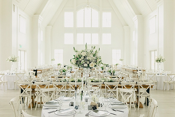 Reception tablescape with a wedding table centerpiece in a tall glass vase, taper candles, and place settings in a bright white hall with chandelier