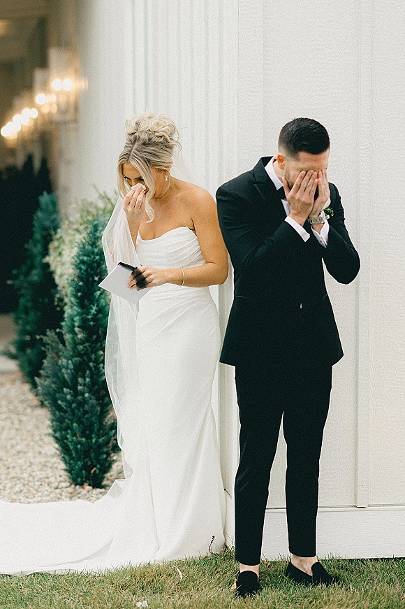 First look moment as bride in strapless dress reads a vow letter while groom in tux wipes tears by a white barn wall with bistro lights