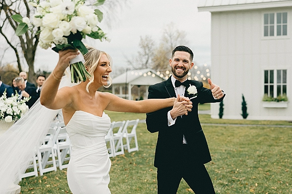 Wedding recessional as bride lifts her bouquet and laughs, holding hands with groom in black tux under string lights on a lawn aisle