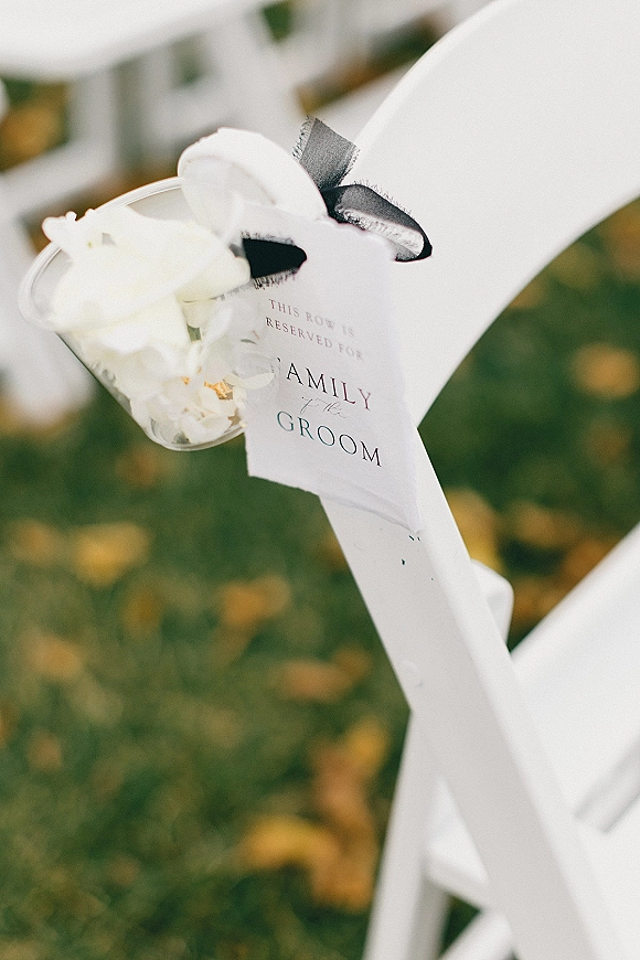 Reserved seating sign tied with black ribbon and a white floral corsage on a white folding chair on a grass lawn with scattered leaves