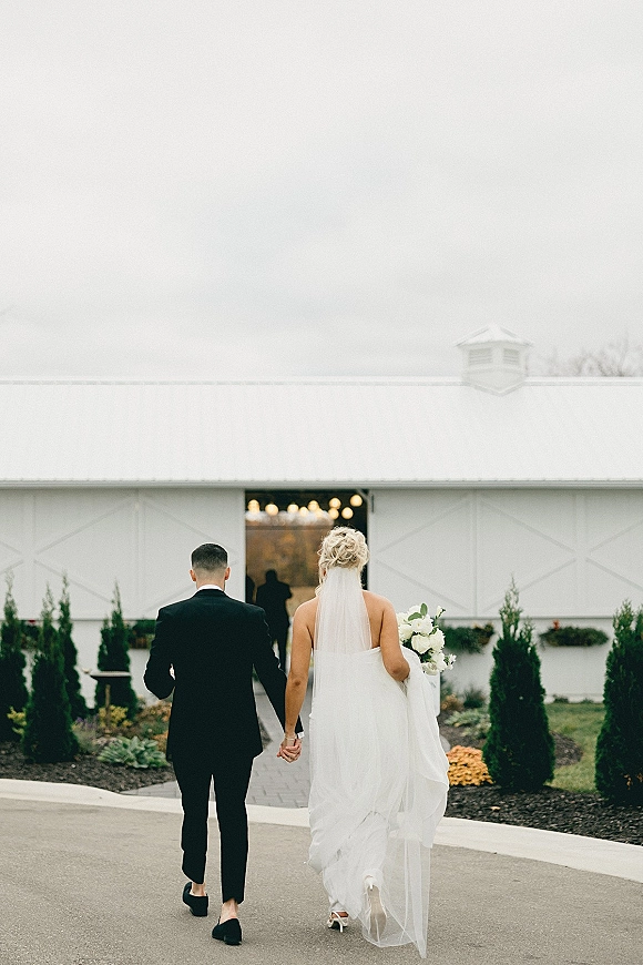 Newlywed couple walking hand in hand toward a white barn entrance, bride in veil holding a bouquet under string lights on a paved path