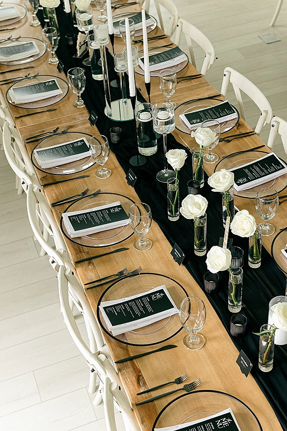 Reception tablescape with long banquet table decor featuring a black table runner, clear chargers, white roses, and tall taper candles on white tiles