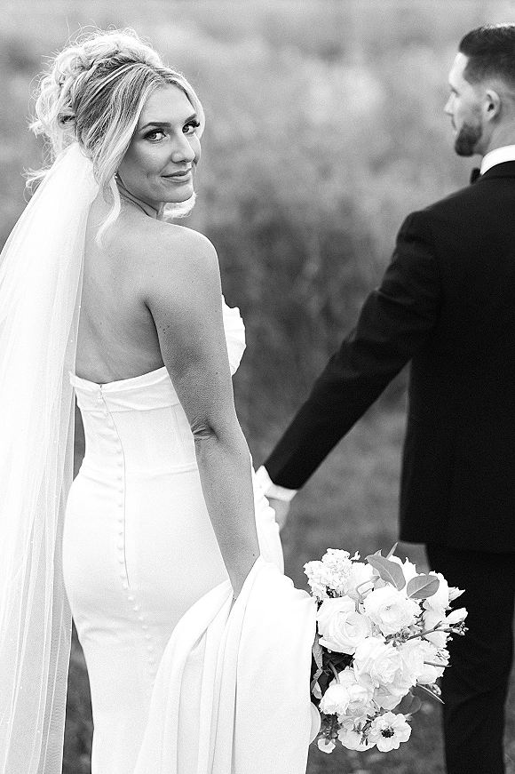 Couple portrait in a black and white wedding portrait, bride looking back holding groom’s hand in a soft-focus outdoor field with veil
