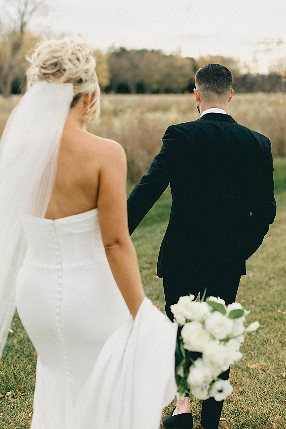 Wedding first look as the bride approaching groom in a grassy field, seen from behind with long veil and white rose bouquet