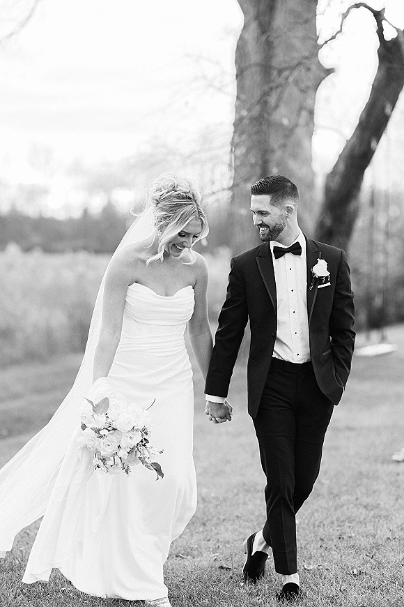 Couple portrait of bride and groom holding hands, laughing as they walk on a grassy lawn with trees, bride in veil and white bouquet
