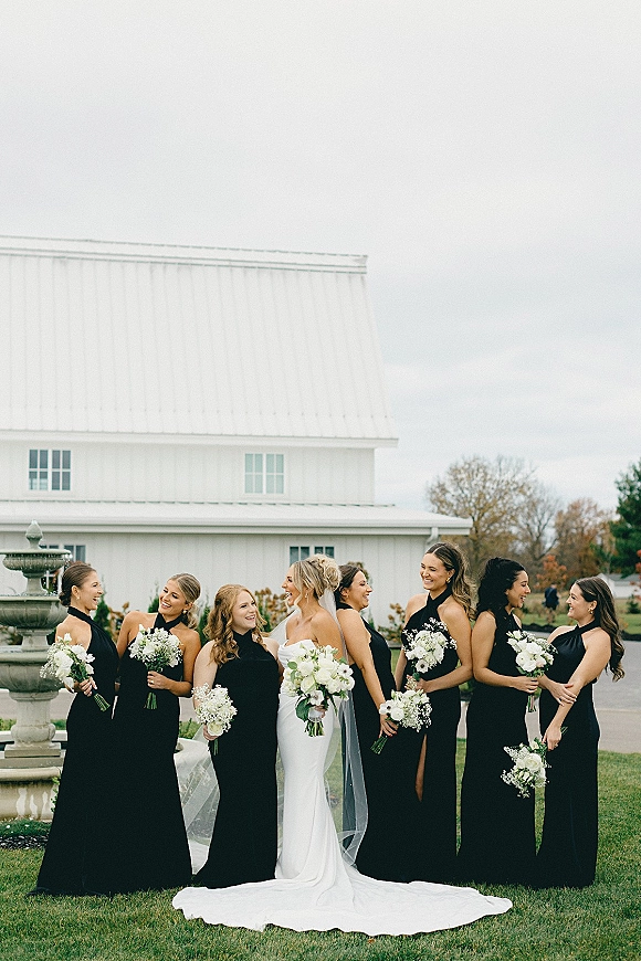 Bridesmaid group photo with bride in veil and strapless dress, bridesmaids in black dresses holding white rose bouquets by a fountain barn lawn
