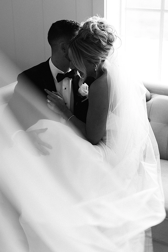 Wedding couple portrait in black and white with bride in veil leaning into groom in tuxedo on a sofa, softly lit by window light indoors