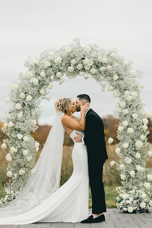 Wedding kiss portrait of bride and groom kissing beneath a white rose floral arch, her veil flowing over stone pavers in an overcast field
