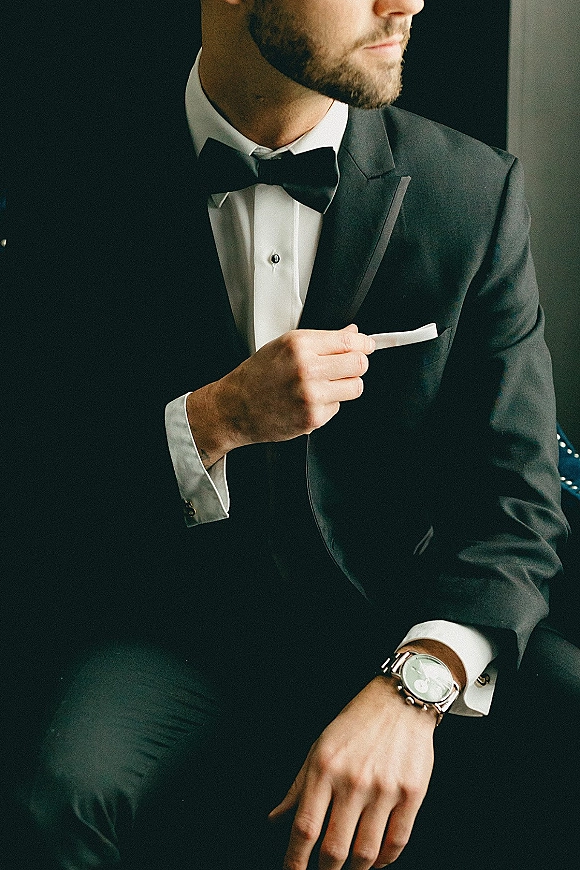 Groom portrait in a groom tuxedo, seated in a dark interior, wearing a black tuxedo with bow tie, white pocket square, and wristwatch